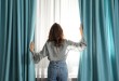 Woman opening window curtains at home in morning, back view