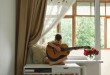 Cute boy learns to play the classical guitar on the windowsill near the window.