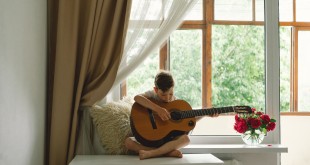 Cute boy learns to play the classical guitar on the windowsill near the window.